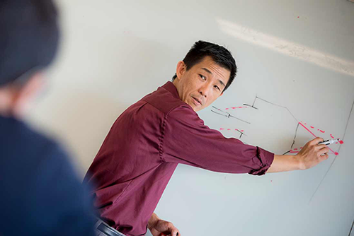 An asian man in a purple shirt points to a diagram on a whiteboard.